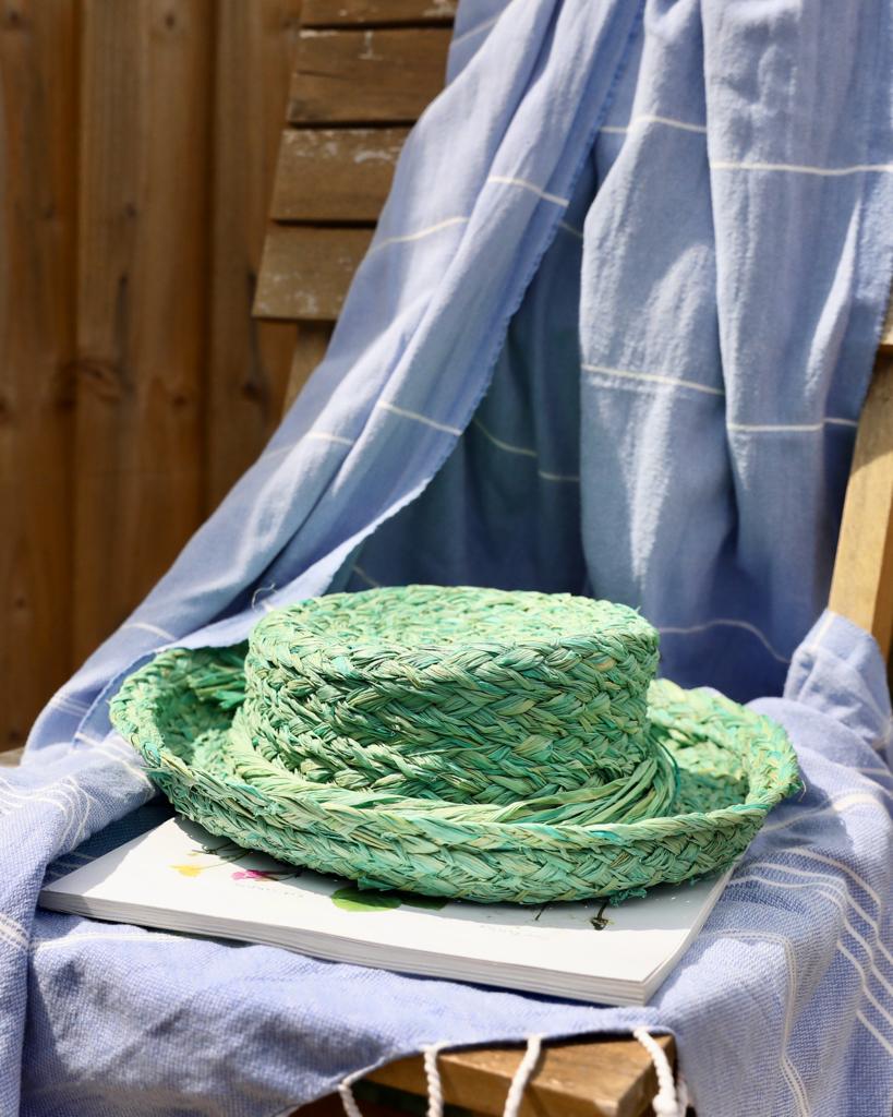 Light green handmade raffia hat placed on a magazine and blue towel in sunshine in garden.  Can be created using complete beginners raffia course