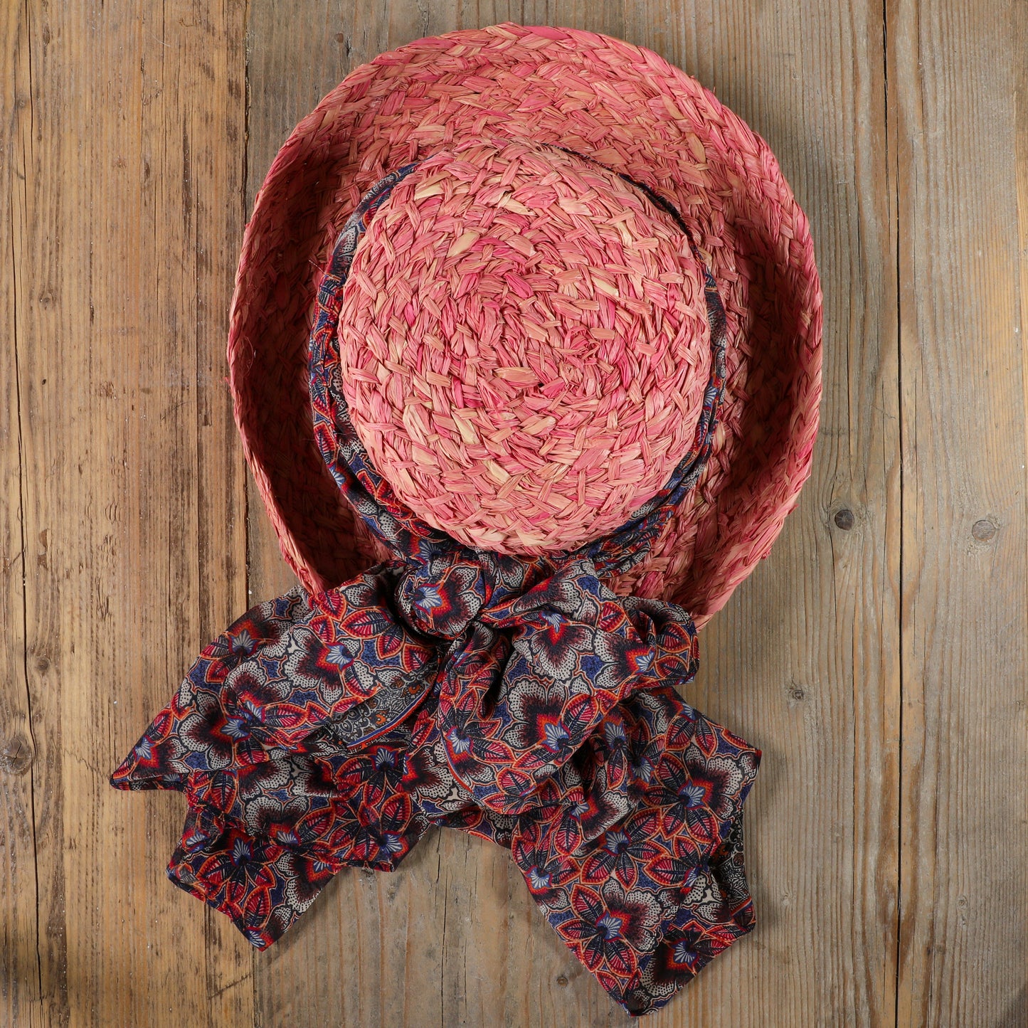 Pink dyed handmade raffia hat with upturned brim on wooden surface with a colourful scarf tied around brim