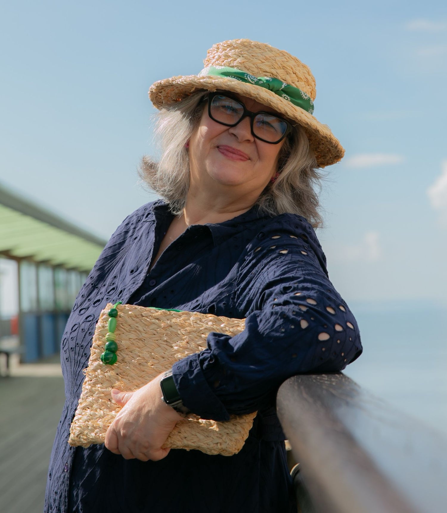 Model with grey hair and glasses navy dress wearing handmade raffia hat with green decor and holding clutch bag with green beads on UK seaside pier