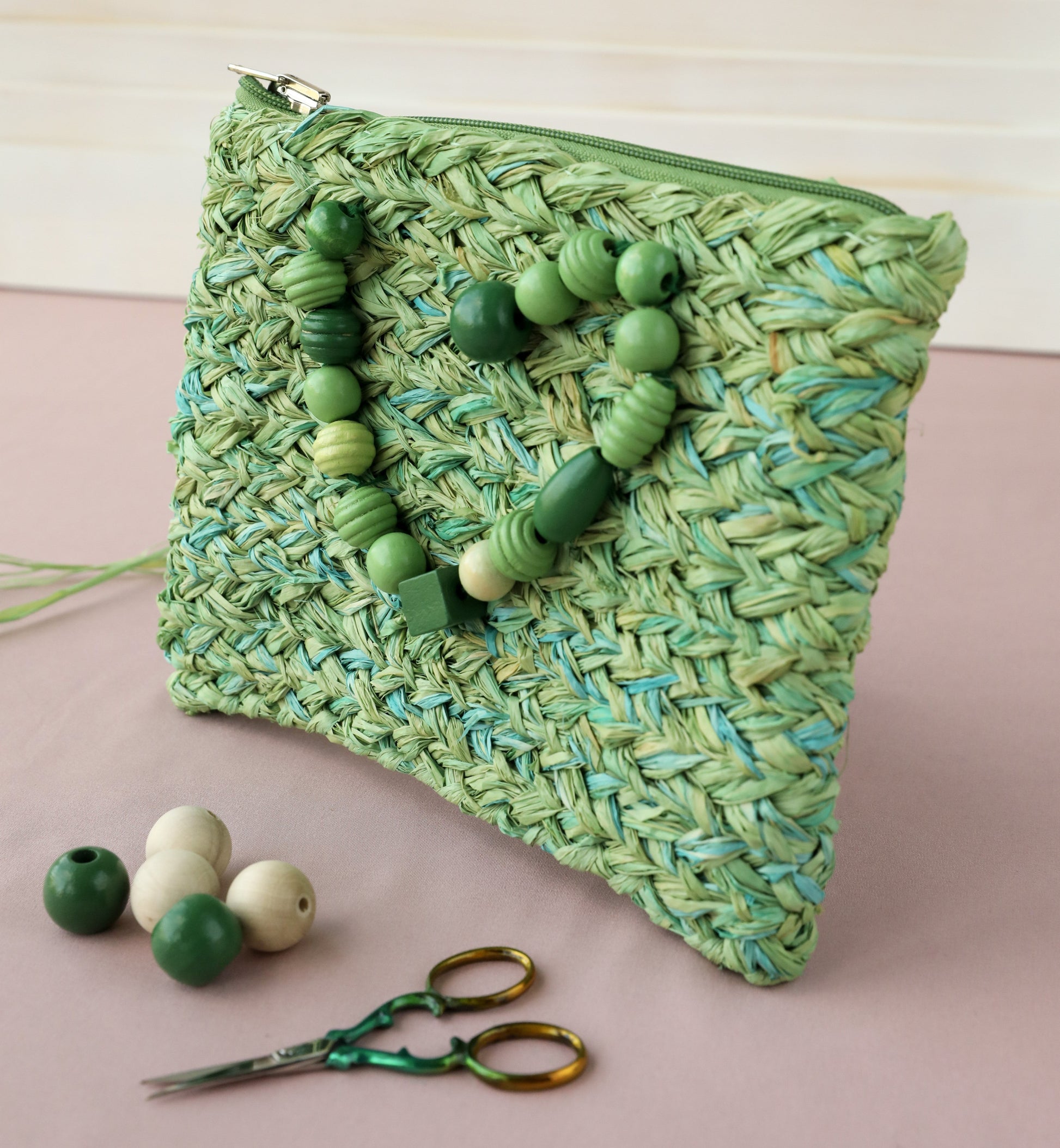 Green raffia  A5 clutch bag with zip and green bead semi-heart detail on pink surface with beads and scissors on table also