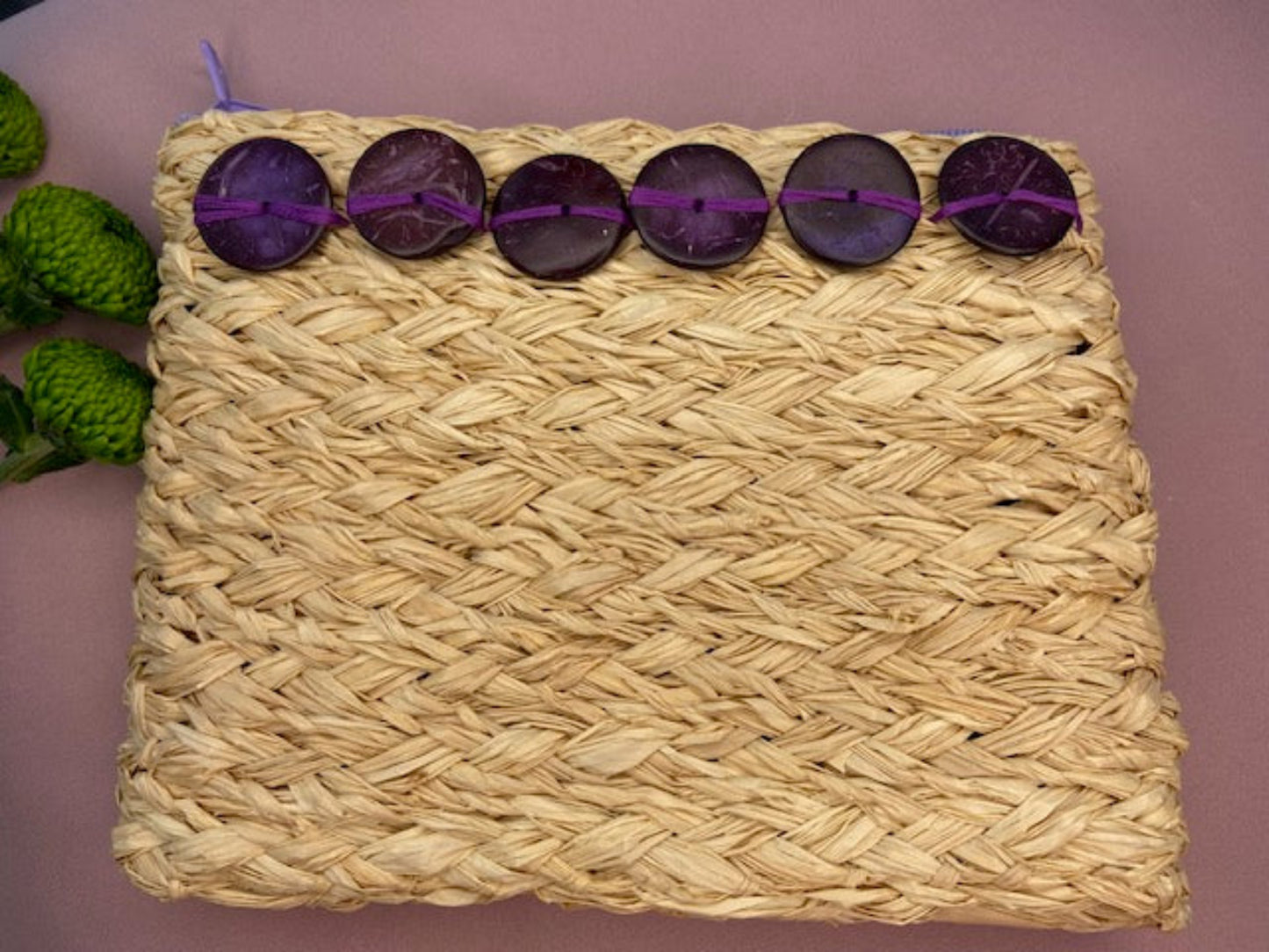 Raffia A5 size clutch bag with purple zip and purple wooden button decoration, on pink surface