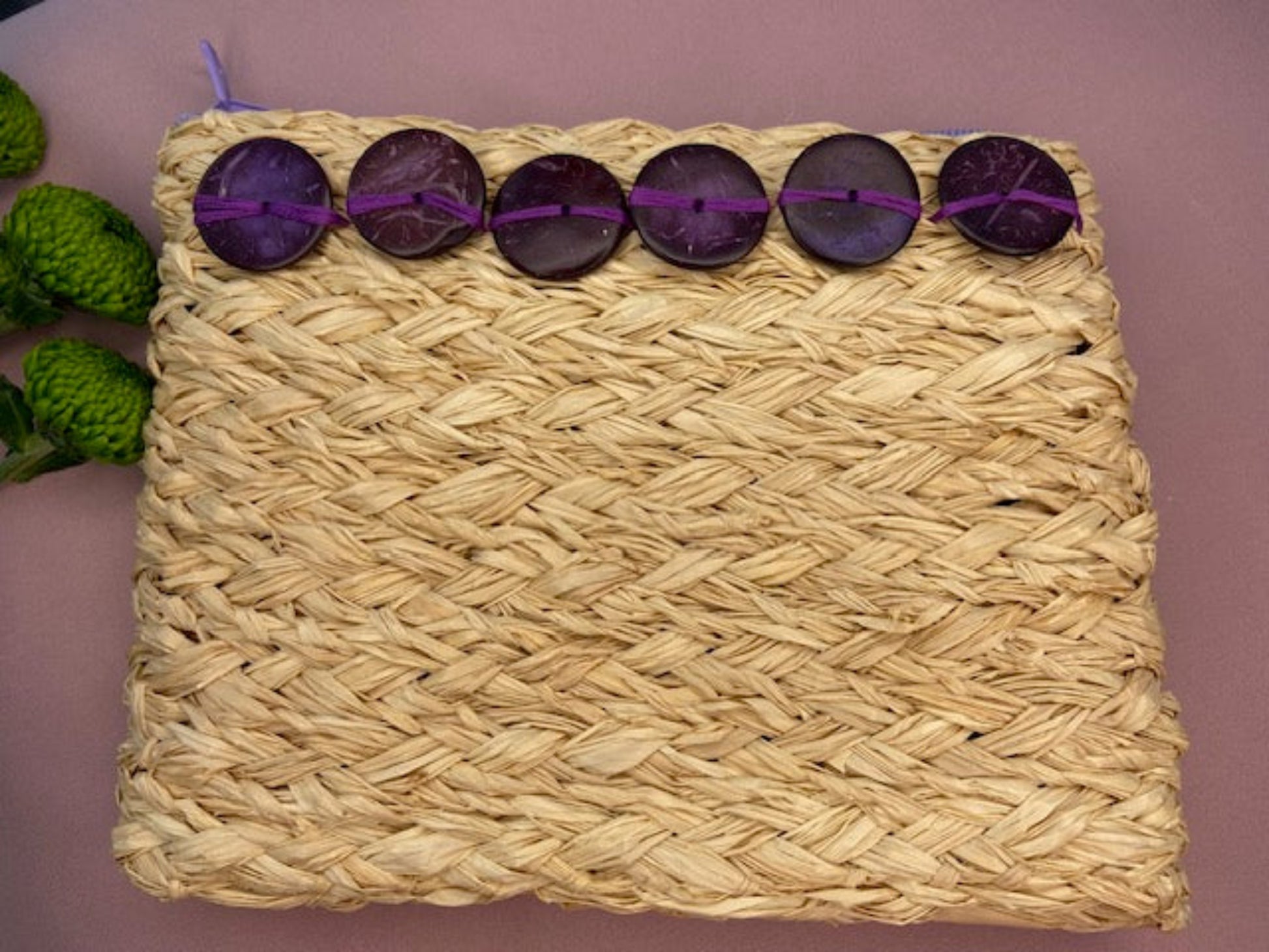 Raffia A5 size clutch bag with purple zip and purple wooden button decoration, on pink surface
