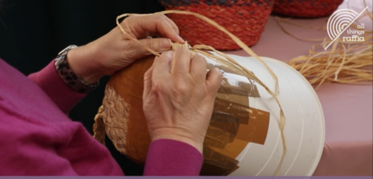 Video screenshot of person sewing a raffia plait on a bucket to create a bag 